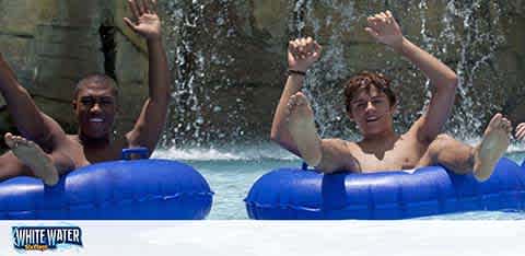 Two people are enjoying a water ride in an amusement park, each sitting in a blue inflatable tube. They are raising their arms in excitement and appear to be smiling as they splash down the water slide, with water cascading in the background. The scene suggests a fun and thrilling experience. The watermark indicates the ride is possibly called 'White Water.'