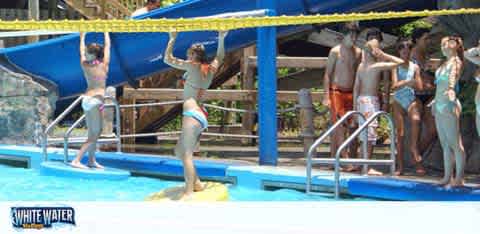 Image shows a group of people at a water park preparing to use a water slide. Individuals in swimwear are standing on a platform beside a blue slide, with some holding onto railings. The water park's name, visible on a sign, implies a fun and wet adventure awaits.