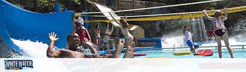 Excited visitors enjoy a sunny day at a water park. A person glides down a water slide in the foreground, while another person in a splash pool raises their arms in delight. To the right, someone stands by a water cannon, preparing to spray the sliders. The park's lively atmosphere is highlighted by splashing water and bright sunlight. Text at the bottom reads 'WHITE WATER'.