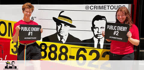 Two individuals stand smiling in front of a stylized 'mugshot' backdrop featuring vintage gangster silhouettes. They each hold signs saying 'PUBLIC ENEMY #1' and 'PUBLIC ENEMY #2' with a hashtag promoting crime tours. The backdrop includes a contact number for the tours.