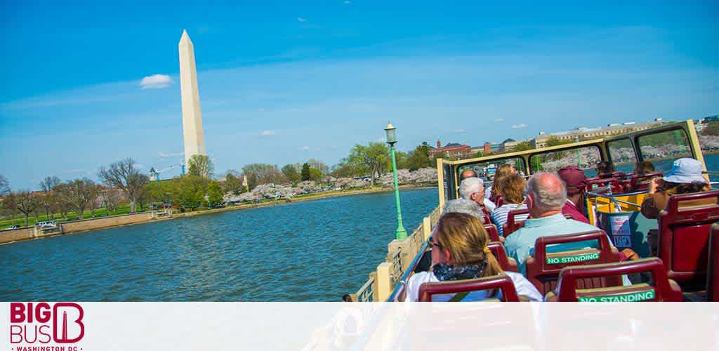 Passengers on a Big Bus sightseeing tour enjoy a sunny day, viewing the iconic Washington Monument across a tranquil expanse of water, with clear skies above and lush greenery along the horizon.