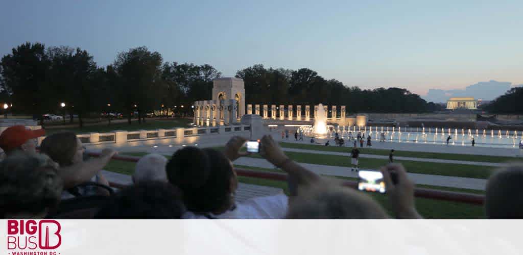 Passengers on a Big Bus tour enjoy a picturesque evening view of a well-lit memorial with fountains. In the foreground, visitors are taking photos while the calm blue sky fades into dusk. The distant memorial is illuminated, creating a serene atmosphere.