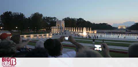 Visitors are viewing a lit monument across a reflecting pool at dusk. Some are capturing the moment with their phones. In the foreground, there's a logo with the text 'Big Bus Tours'. The setting appears peaceful, and the sky is transitioning from blue to soft twilight hues.