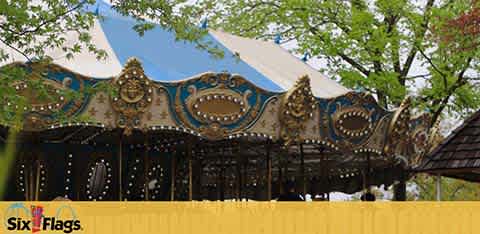 Image shows a carousel under a large, white-tipped blue tent at a Six Flags amusement park. Decorative golden accents and lights adorn the ride, with green trees and a clear sky in the background. The Six Flags logo is visible at the bottom.