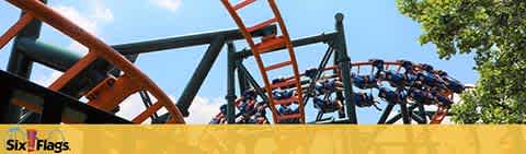 Image shows a roller coaster with orange tracks against a blue sky, filled with people secured in the seats as the ride twists along the track. Trees are visible in the background, and the 'Six Flags' logo is displayed in the corner.