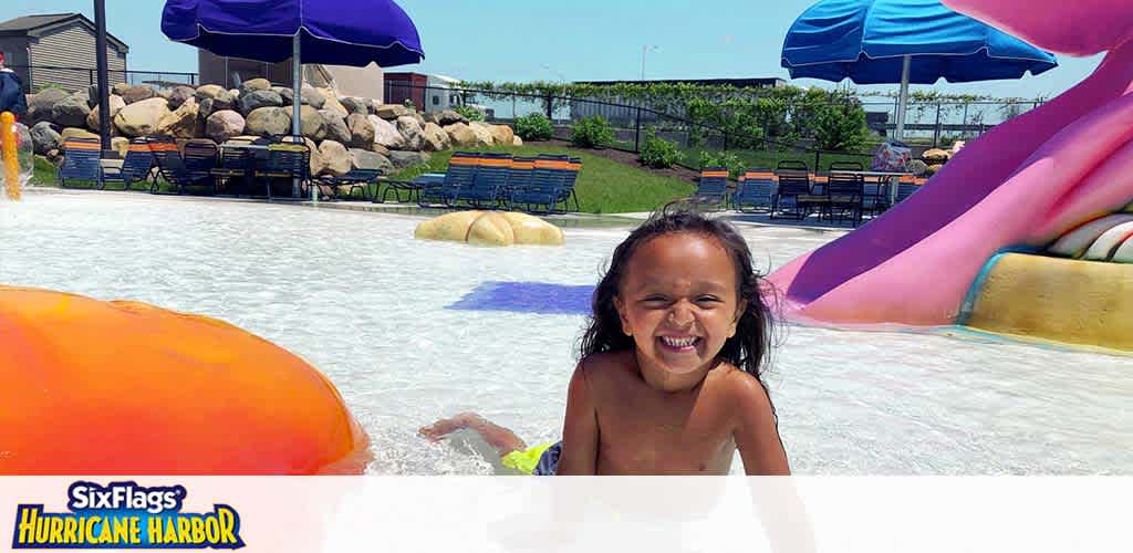 A joyful child is splashing in a shallow water play area on a sunny day at Six Flags Hurricane Harbor. Colorful water slides and umbrellas are in the background, along with lounge chairs for relaxation. Vibrant orange and purple hues add to the fun atmosphere of the waterpark.