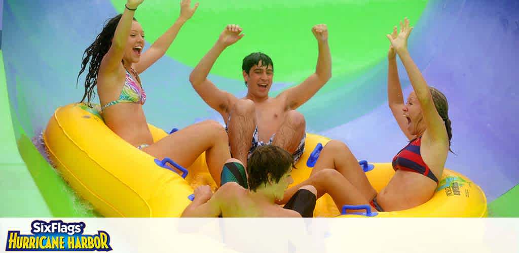 Image shows a group of four excited people in swimwear sliding down a bright, multicolored water slide on a large yellow inflatable raft with handles. The background is a blend of vibrant green and blue, suggesting high speed and water-based fun at Six Flags Hurricane Harbor.