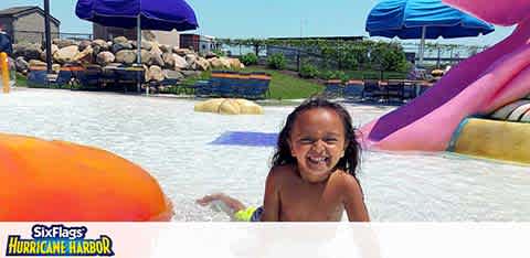 A child with a joyful smile is playing in a shallow water area at Six Flags Hurricane Harbor. In the background purple water slides, blue umbrellas, and rock formations are visible under a clear sky. The park's logo is seen at the bottom.