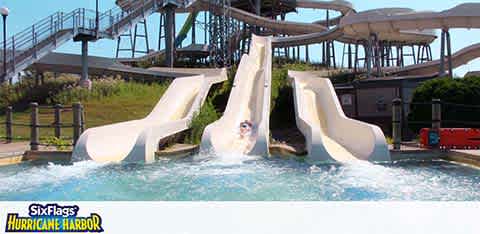 The image shows a bright sunny day at a waterpark, specifically Six Flags Hurricane Harbor. Three parallel water slides with white surfaces and blue edges descend towards a common splash pool. A person is visible on the left slide, enjoying the ride, moments away from the water. The slides are surrounded by a network of walkways and lush greenery in the background.