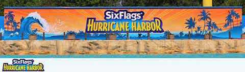 Image of a vibrant promotional banner for Six Flags Hurricane Harbor water park. The banner features a stylized wave and palm tree graphics with the park's name in large, bold letters, set against a backdrop of a tropical blue sky. Below is a reflection of the sign on a water surface. The Six Flags logo is present at both top and bottom left corners.