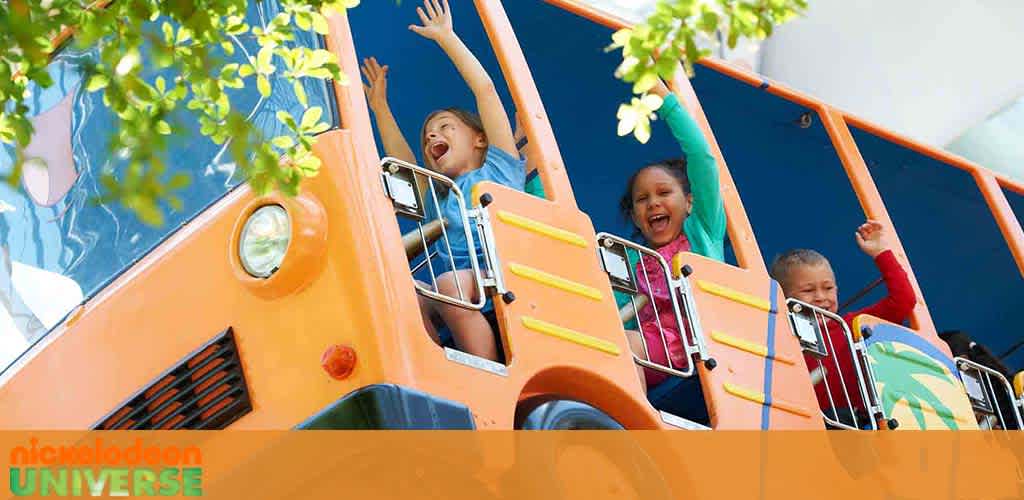 Image shows children with arms raised in excitement on an orange bus-themed amusement park ride. Bright blue skies, green foliage, and the  Nickelodeon Universe  logo add to the joyful atmosphere.