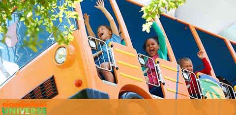The image showcases three joyful children on a bright orange amusement park ride that simulates a bus. The sky is visible in the background with a hint of green foliage at the top left corner. The ride's front resembles a cartoonish bus design with large, round headlights. The children have their hands raised in excitement, and the atmosphere conveys a sense of fun and adventure.