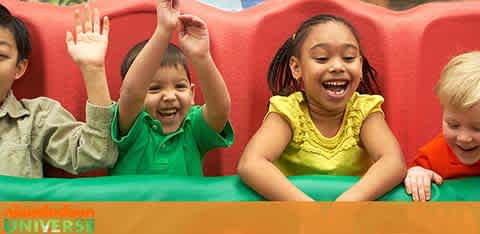 Image of four joyful children sitting in a row on a colorful playground structure. They are variously raising their hands and laughing, expressing delight. Colors are vibrant with red, green, and orange dominating the scene. The children appear to be of diverse ethnic backgrounds, sharing a happy moment together.