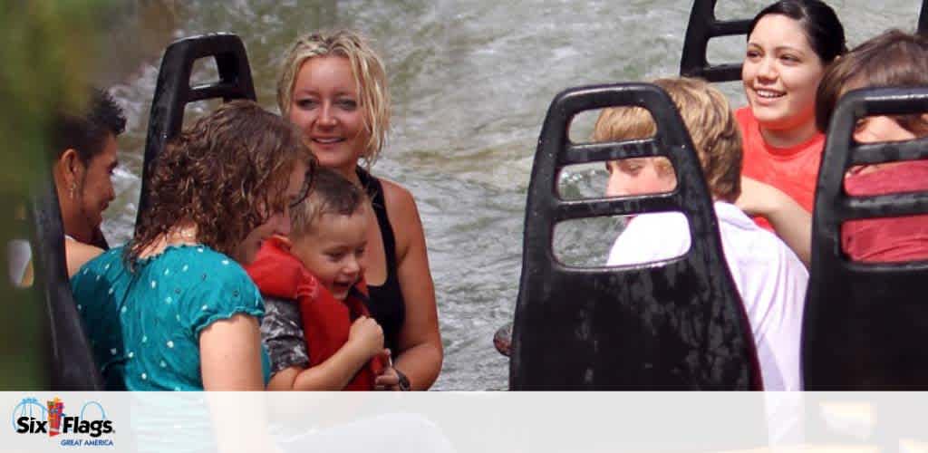 Guests enjoy a water ride at an amusement park, smiling with excitement. Some are drenched in water, capturing the fun of a splashy adventure. The Six Flags logo indicates the park's branding. A sense of joy and amusement pervades the scene.