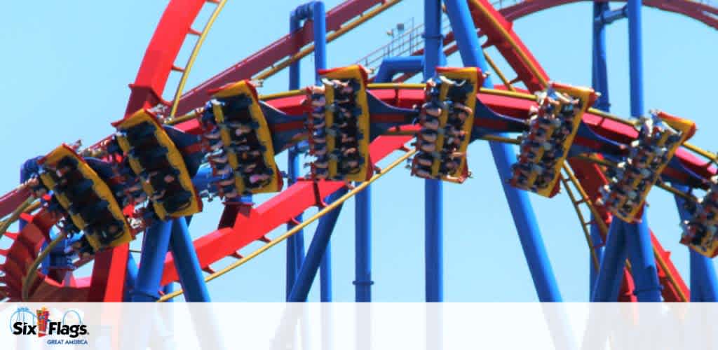 Image shows a roller coaster with red tracks and blue supports at a Six Flags theme park. Thrilled riders in multiple cars are inverted on a loop, against a clear blue sky. The Six Flags logo is visible, indicating the location is Great America.