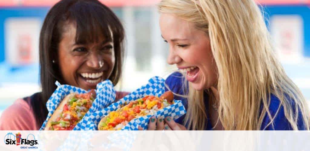 Two joyful women are holding and about to eat large, fully-loaded hot dogs wrapped in checkered paper at an amusement park. The woman on the left, with dark hair, smiles broadly, while the blonde woman on the right laughs with delight. A logo at the bottom indicates the location is Six Flags Great America. The scene exudes a vibrant, fun atmosphere.