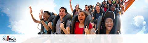 Image of a diverse group of people on a roller coaster ride at Six Flags amusement park. They are seated in rows, with hands raised, expressing thrill and excitement. The sky is clear and the Six Flags logo is seen in the lower right corner.