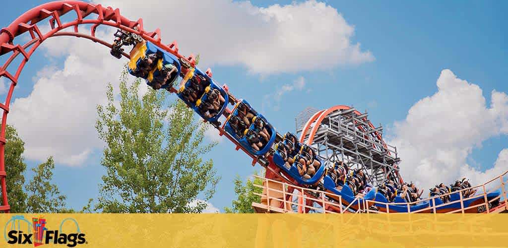 Image of a roller coaster with a red track and blue carriages filled with riders in harnesses, upside-down as they navigate a loop. Lush green trees, a blue sky with wispy clouds, and the logo for Six Flags amusement park are also visible.