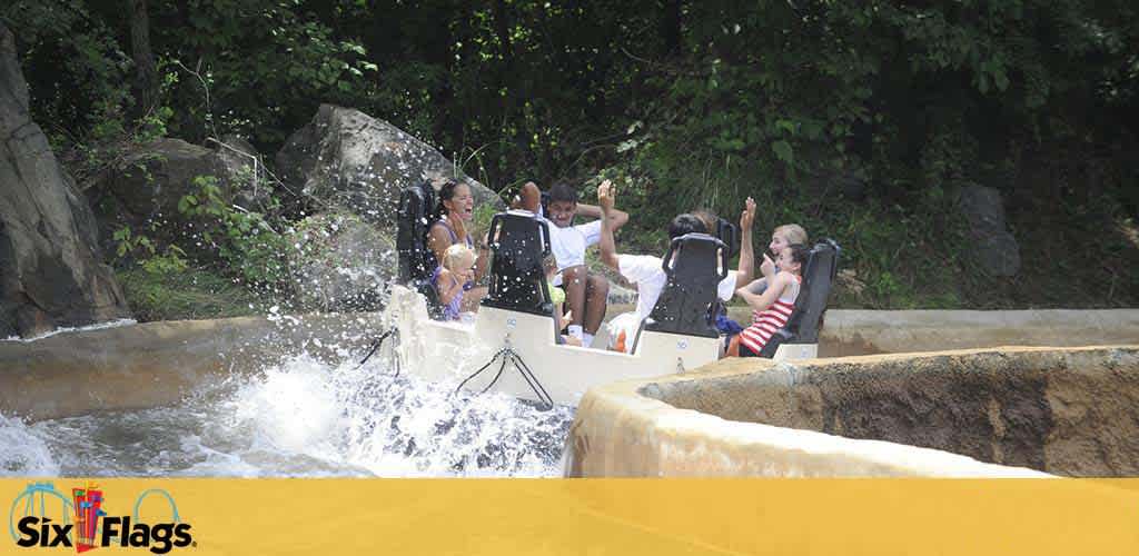 Visitors enjoy a water ride at Six Flags, splashing down a flume surrounded by rocks and trees. Excitement is evident as water sprays around their raft on a sunny day.