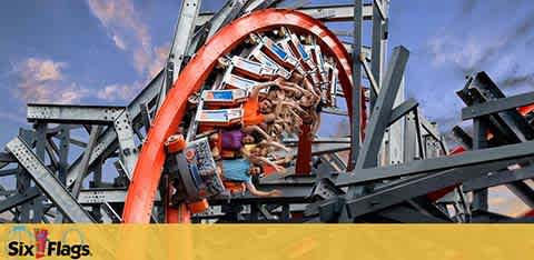 Image depicts a roller coaster with a red track and grey support structure, where riders in multiple rows are experiencing an inverted loop. They appear thrilled. A blue sky with light clouds forms the background, and the Six Flags logo is seen in the corner on a yellow stripe.
