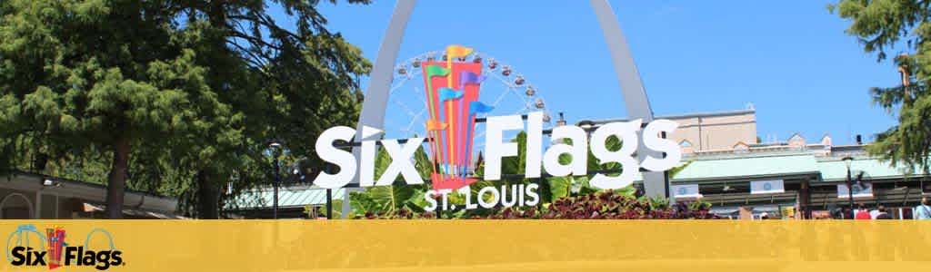 Image of the entrance sign for Six Flags in St. Louis on a clear day. The iconic logo is white against a blue sky, flanked by trees and colorful roller coaster tracks in the background. In the foreground, a yellow barrier with greenery shows the park's vibrant atmosphere.