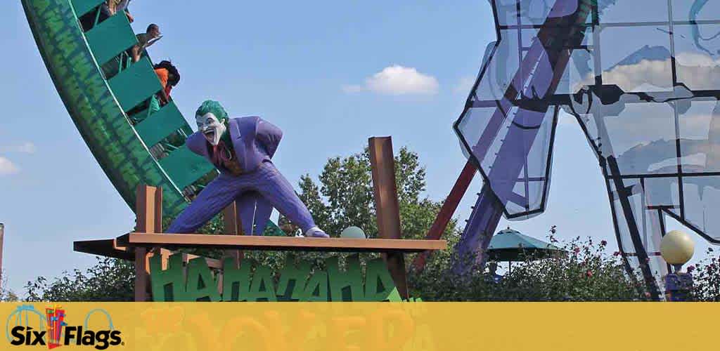 Image shows a dynamic amusement park scene at Six Flags featuring a large, colorful statue of an animated character who resembles the Joker, laughing and positioned in front of a roller coaster filled with riders. The coaster is in a visceral banked turn, enhancing the excitement. The theme park's logo is visible in the foreground. Skies are clear with scattered clouds, suggesting a lively day out.