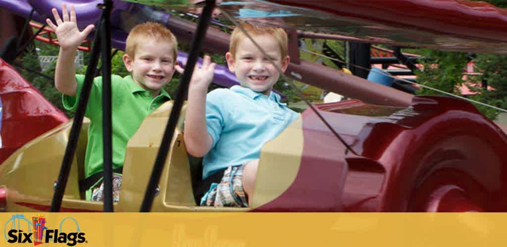 Two joyful children are enjoying a ride in an amusement park, sitting in a car-like structure with a reddish-brown front. The boy on the left, in a green shirt, raises his hand high, while the boy on the right, in a light blue shirt, waves delightedly. In the background, blurred foliage and park structures suggest a cheerful, bustling atmosphere. The Six Flags logo is visible, indicating the location.