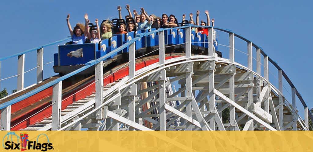 A group of excited riders raises their arms in sheer joy on a blue roller coaster cart descending a steep wooden track against a clear blue sky. Below the track, there is a yellow barrier and the Six Flags logo is visible, suggesting the setting is a theme park.
