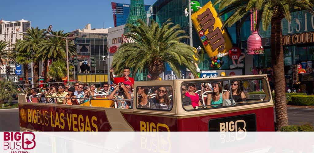 Image shows a Big Bus Las Vegas tour with people seated on the open-top deck. Palm trees line the street with vibrant commercial buildings in the background, under a clear blue sky.