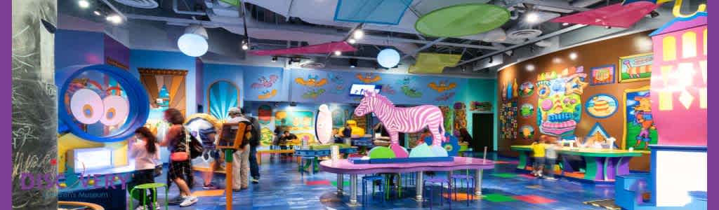 Colorful indoor play area with vibrant walls, floor, and decorations. Features include a large, stripe-painted zebra figure, cartoon-style wall art, interactive exhibits, and tables for activities. People, possibly families, are engaging with the exhibits and each other under bright lighting.