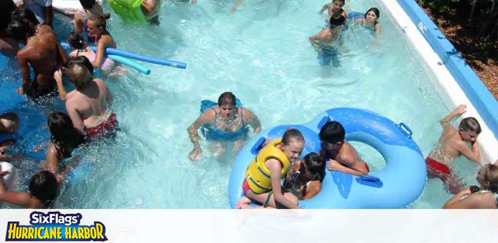 The image displays a lively scene at a water park with multiple visitors enjoying themselves in a pool. Some individuals are floating on rafts while others are swimming or playing with foam noodles. The atmosphere is playful and energetic, with clear blue water, and the watermark indicates the location is Six Flags Hurricane Harbor.