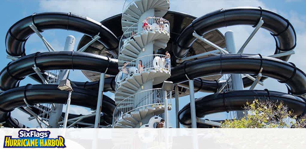 Image shows a towering water slide at Six Flags Hurricane Harbor. Multiple black tubes spiral downward around a central structure with stairs where guests are ascending to the slide entrances. The sky is clear indicating favorable weather for water activities.