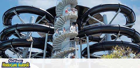 Image of a multi-level water slide structure with a series of intertwined black slides spiraling down around central towers against a blue sky. The base is partly obscured by greenery. Text at the bottom displays Six Flags Hurricane Harbor.