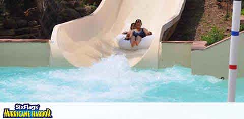 A person is enjoying a water slide at Six Flags Hurricane Harbor. They appear thrilled, descending quickly with water splashing around. The slide is white, curving into a large splash pool with clear, blue water. Green foliage surrounds the scene, adding a natural touch to the amusement park excitement.