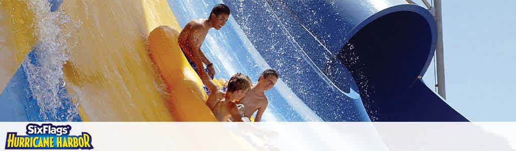 Banner image featuring three individuals having fun on a blue and yellow water slide at Six Flags Hurricane Harbor. Splashes of water can be seen around as they enjoy the ride under a clear blue sky. The Six Flags Hurricane Harbor logo is visible in the corner.