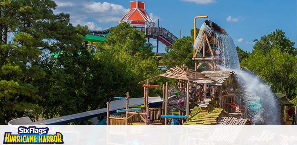 Image shows a vibrant water park with clear skies overhead. A large bucket tips, cascading water onto a wooden play structure surrounded by lush green trees. A green and white slide weaves in the background. The Six Flags Hurricane Harbor logo is displayed at the bottom.