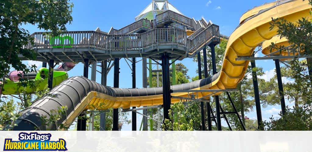 Image of a sunny day at Six Flags Hurricane Harbor showing a large water slide with yellow and black tubes that twist and turn, protruding from a multi-level platform with green railing surrounded by lush trees. The park's logo is visible at the bottom left.