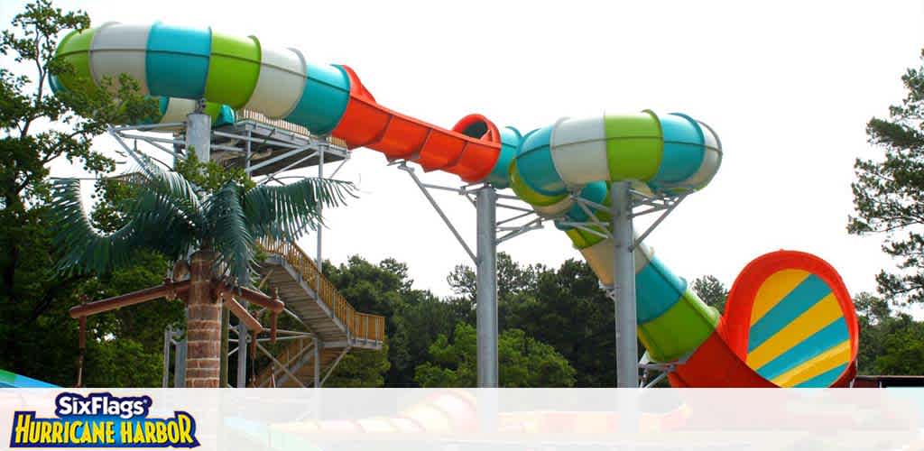 This image shows a vibrant water slide at Six Flags Hurricane Harbor. The slide features a combination of blue, green, orange, and yellow colors with a spiraling design. A faux palm tree decorates the base, and the structure stands against a backdrop of trees under a clear sky.