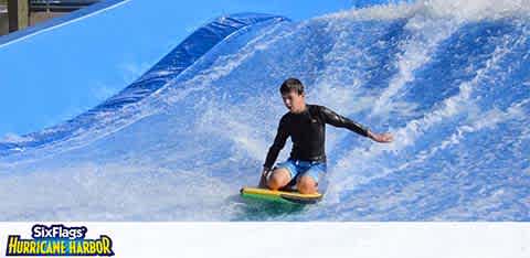 A person in a black and blue wetsuit is surfing on a simulated wave at a water attraction. Splashes of white water envelop the surfer who balances on a yellow board. The logo for Six Flags Hurricane Harbor is visible in the foreground. The sky is clear blue, suggesting a sunny day.