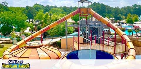 Image depicts a vibrant water slide at Six Flags Hurricane Harbor. In the foreground, a circular pool rests at the base of an orange slide. A multi-level play structure with water features is visible in the middle. Lush green trees encircle the park under a blue sky.