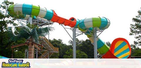 This image features colorful water slides at Six Flags Hurricane Harbor. The slides are a mix of enclosed tubes with stripes and open-air slides with spirals. Trees are visible in the background. The sky is overcast and the park's logo is included in the frame.