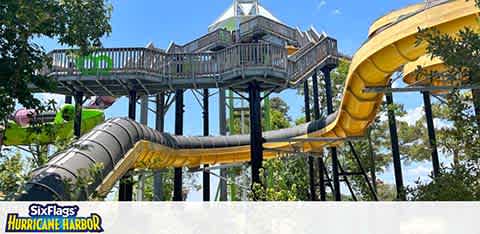 Image of a water slide at Six Flags Hurricane Harbor featuring a towering structure with multiple slides. One slide is yellow with open and enclosed sections amidst lush green trees under a clear blue sky. The park's logo is visible in the foreground.