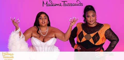 Image features two figures against a pink background, associated with Madame Tussauds. On the left, a figure in a white dress with fluffy sleeves poses with hands up. To the right, another figure wears a black outfit with orange and beige patterns, one hand on the hip. Both figures are wax representations and appear lifelike.