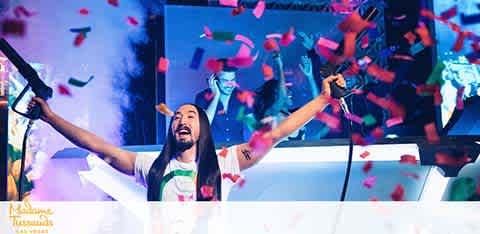 An energetic DJ with long hair is cheering on stage, hands raised high amidst a burst of colored confetti. Bright lights and visuals enhance the vibrant atmosphere of the music festival.