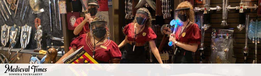 Image shows three people in a Medieval Times gift shop. They're wearing red tunics with emblems and facemasks. One is trying on a knight helmet, while another holds a blue sword-like toy. The background is filled with medieval-themed merchandise. The Medieval Times logo is visible at the bottom.