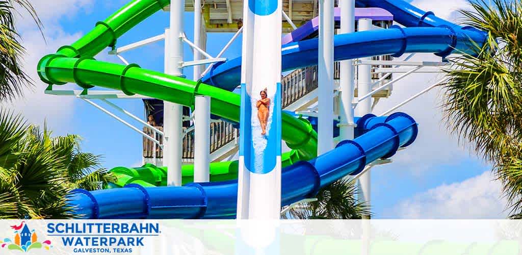 Photo of a vibrant waterpark at Schlitterbahn in Galveston, Texas. Bright blue and green water slides twist around white support structures against a backdrop of tropical palm trees and a clear sky. A person is visible sliding down, enjoying the excitement. The park logo is in the bottom left corner.