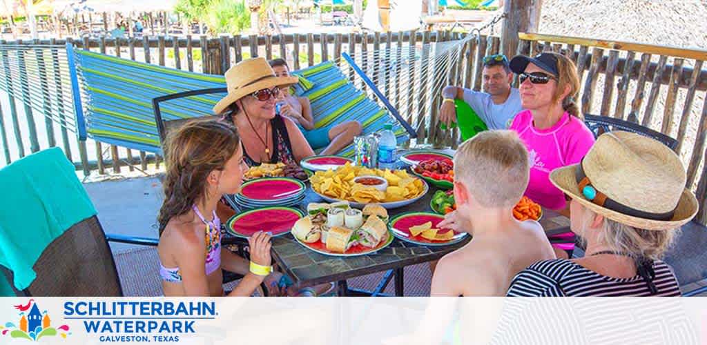 A group of guests enjoying a meal at Schlitterbahn Waterpark in Galveston, Texas. They are seated around a picnic table filled with colorful plates and a variety of food under a thatched-roof cabana. The atmosphere is casual and sunny with visible palm trees, suggesting a relaxed, tropical setting.