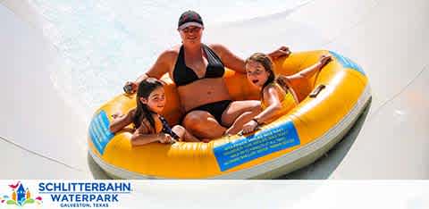 Image depicts three joyful individuals on a yellow water raft sliding down a water slide with a sign indicating 'Schlitterbahn Waterpark Galveston, Texas' in the background, capturing a moment of fun and excitement at the waterpark.