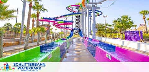 Image shows Schlitterbahn Waterpark in Galveston, Texas, with a view of a vibrant, multi-lane water slide. Brightly colored in pink, purple, and blue, the slides twist and turn above a spacious pool area. Clear skies and palms frame the scene, inviting visitors to a day of water fun.