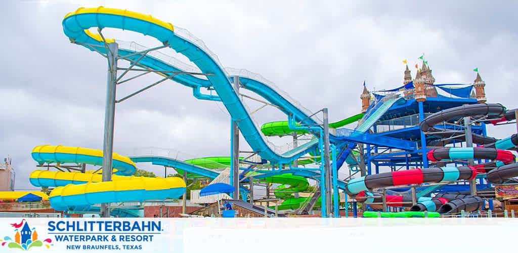 Image shows colorful water slides at Schlitterbahn Waterpark and Resort in New Braunfels, Texas. Twisting slides in bright blue and green hues with multi-person rafts ready at the start platforms. In the background, a castle-like structure with turrets under a cloudy sky.